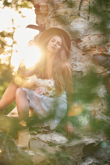 Impresionante joven con un vestido blanco de verano y un sombrero con peonías blancas en sus manos