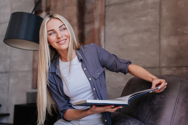 Impresionante modelo rubia relajándose en casa sentada con un libro en una silla acogedora mira a un lado felizmente