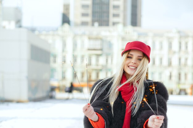 Impresionante modelo rubia sonriente con gorra de moda divirtiéndose con bengalas en la calle. Espacio para texto