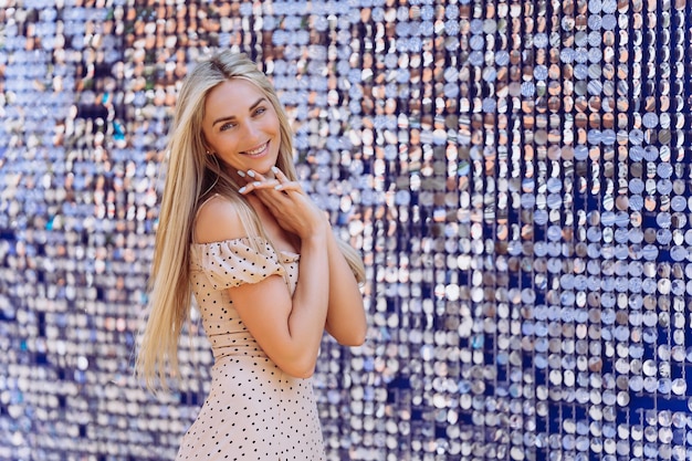 Impresionante mujer joven rubia con cabello largo suelto en vestido beige doblando las manos en el pecho con una sonrisa adorable mirando a la cámara contra un fondo brillante con espacio vacío Concepto de moda y compras