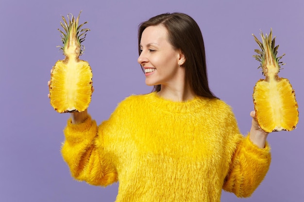 Impresionante mujer joven en suéter de piel mirando a un lado sosteniendo mitades de fruta fresca de piña madura aislada sobre fondo violeta pastel. Estilo de vida vivo de la gente, concepto de vacaciones relajantes. Simulacros de espacio de copia.