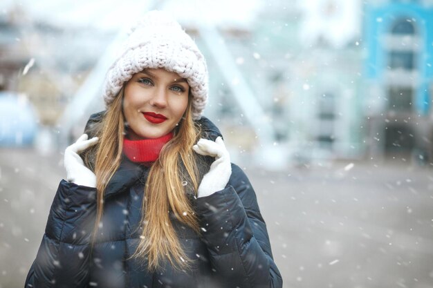 Impresionante mujer rubia con gorro tejido caminando en la ciudad durante las nevadas. Espacio para texto