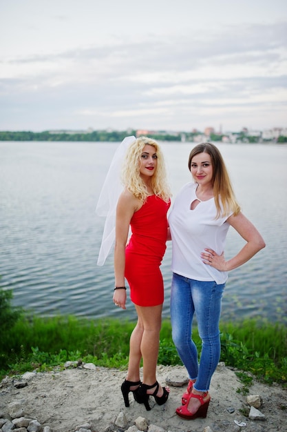 Impresionante novia en vestido rojo posando con su dama de honor en la orilla del lago