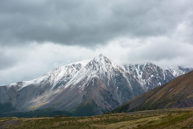 Impresionante paisaje atmosférico con un alto pico de montaña nevado iluminado por el sol en nubes bajas lluviosas en un clima cambiante Dramática hermosa vista desde la colina dorada hasta la gran cima de la montaña nevada en un cielo nublado gris bajo