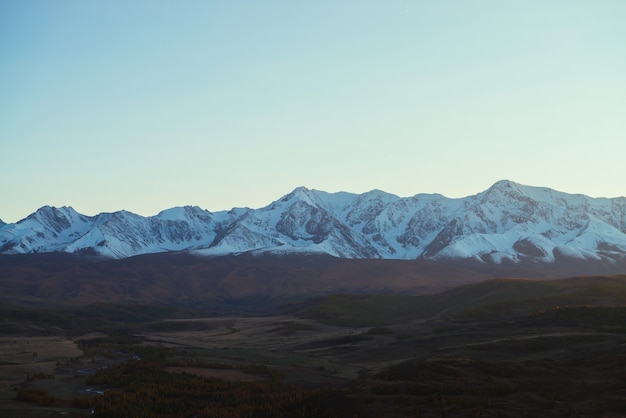 Impresionante paisaje con valle de montaña con colinas y bosque en colores otoñales a la sombra con vista a la gran cordillera nevada en la puesta de sol. Altas montañas cubiertas de nieve y valle otoñal con poca iluminación