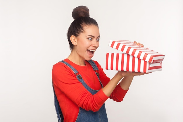 Impresionante regalo de vacaciones. Retrato de una chica guapa entrometida sorprendida con moño de pelo en overoles de mezclilla mirando a la caja actual con expresión curiosa y sorprendida, conmocionada por lo que hay dentro. foto de estudio