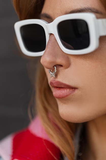 Impresionante retrato de primer plano de la calle de una mujer bonita de la moda con elegantes gafas de sol blancas con ruido penetrante en ropa urbana de moda sobre un fondo oscuro Cara hipster femenina