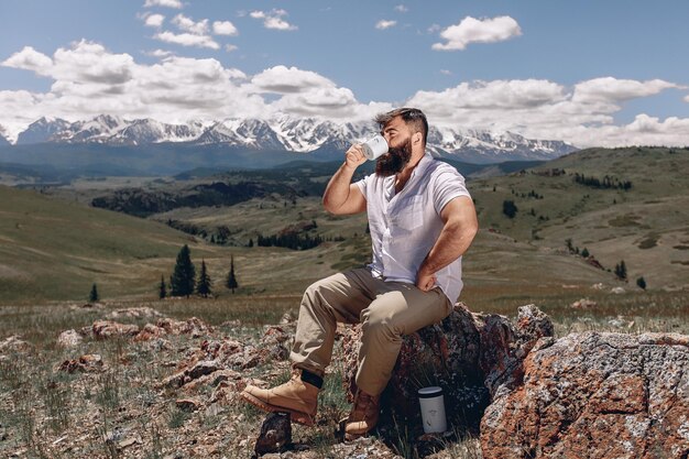 Impresionante y tranquilo paisaje de montaña. Un hombre adulto con barba en una camisa blanca está sentado en una piedra en un césped verde y bebiendo té caliente. Hay un termo blanco debajo del adoquín.