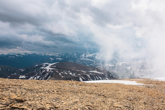 Impresionante vista superior a través de las nubes a altas montañas nevadas Paisaje escénico con hermosas montañas nevadas en nubes bajas Vista alpina atmosférica desde la colina de piedra hasta la cordillera nevada con nubes bajas