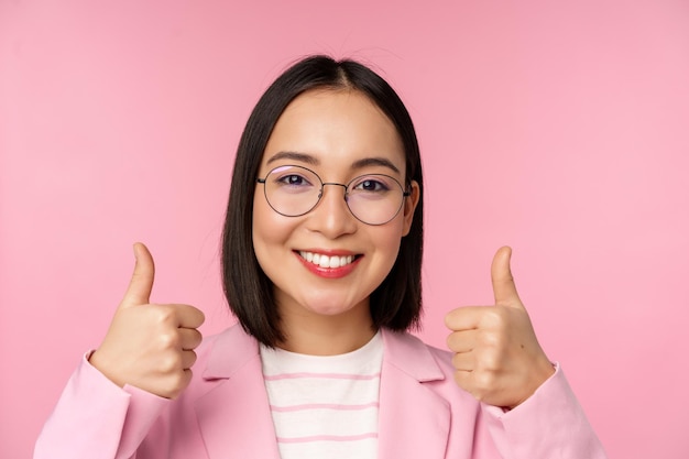 Impresionantes felicitaciones Cara de emocionada mujer de negocios asiática con gafas sonriendo complacida mostrando los pulgares hacia arriba en señal de aprobación de pie sobre fondo rosa