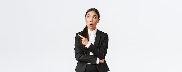 Impressionada empresária gerente feminina bastante asiática de terno apontando e olhando o canto superior esquerdo com expressão espantada local bom negócio em pé fundo branco Copiar espaço