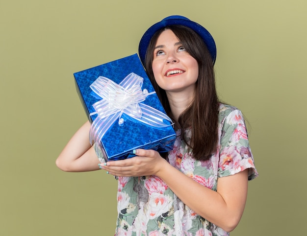 Impressionado ao olhar para uma jovem linda com um chapéu de festa segurando uma caixa de presente ao redor do rosto, isolada na parede verde oliva