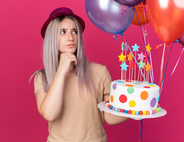 Impressionado ao olhar para uma jovem linda usando um chapéu de festa segurando balões com um bolo agarrado no queixo isolado na parede rosa