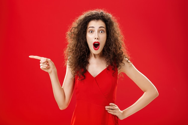 Impressionado até o fundo do coração. Retrato de mulher bonita, intrigada e surpresa, em um vestido de noite vermelho elegante, apontando para a esquerda, divertida e espantada, deixando cair o queixo de espanto.