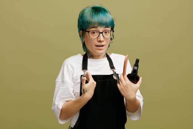 Impressionado jovem barbeiro feminino vestindo uniforme de óculos segurando pentes e spray de cabelo olhando para a câmera apontando para si mesma com as mãos isoladas no fundo verde oliva