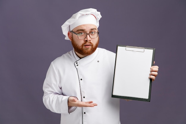 Impressionado jovem chef vestindo óculos uniforme e boné olhando para a câmera mostrando a área de transferência apontando para ele com a mão isolada no fundo roxo