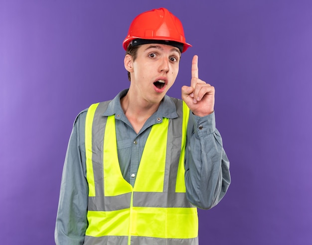 Impressionado olhando para a câmera, o jovem construtor de uniforme aponta para cima