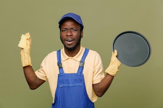 Impressionado segurando a bandeja com esponja jovem limpador afro-americano de uniforme com luvas isoladas em fundo verde