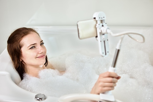 In der Badewanne liegende Frau macht Videoanrufe, indem sie das Telefon in der Halterung befestigt