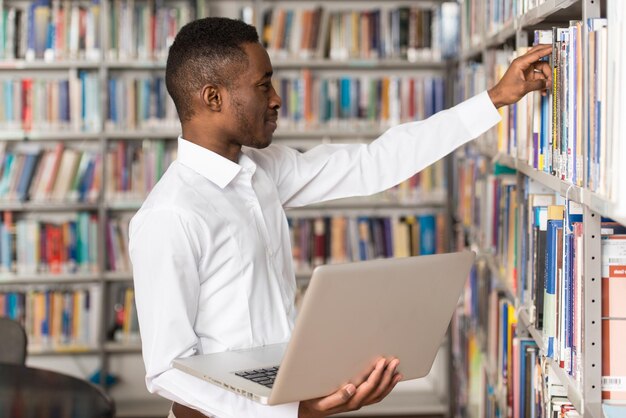 In der Bibliothek Hübscher männlicher Student mit Laptop und Büchern, die in einer High School-Universitätsbibliothek arbeiten Geringe Schärfentiefe