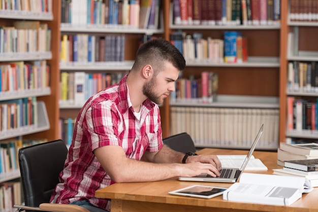 In der Bibliothek Hübscher männlicher Student mit Laptop und Büchern, die in einer High School-Universitätsbibliothek arbeiten Geringe Schärfentiefe