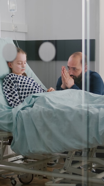 In der Krankenstation schläft ein kranker Mädchenpatient, während der besorgte Vater für die Genesung betet