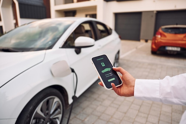 In der Nähe einer Garage Nahaufnahme Frau lädt ihr Elektroauto und überwacht den Vorgang auf dem Smartphone