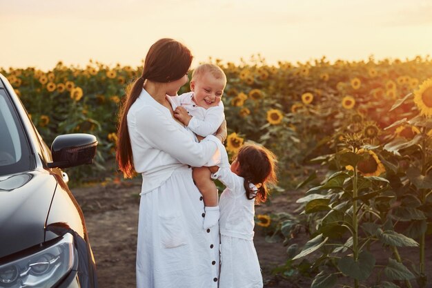 In der Nähe eines modernen schwarzen Autos Junge Mutter mit ihrem kleinen Sohn und ihrer Tochter ist draußen auf dem landwirtschaftlichen Gebiet Schöner Sonnenschein