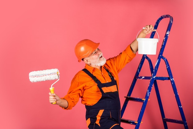 In der Reparaturwerkstatt arbeitender Senior-Maler verwendet eine Walze auf der Leiter, die die Wand in rosa streicht. Professioneller Maler in Arbeitskleidung