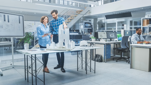 In einem Architekturbüro sprechen Architekt und Ingenieur, die an einem Prototypenprojekt für ein Gebäudekomplex arbeiten, wobei sie Stadtmodelle und Computer verwenden, 3D-CAD-Software betreiben, Wohn- oder Geschäftsviertel