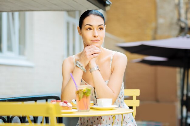 In einem Cafe. Ruhige schöne junge Frau, die bequem am Tisch auf einer Caféterrasse sitzt und in die Ferne schaut, während sie auf einen Freund wartet