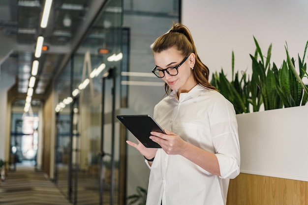 In einem luftigen Korridor untersucht eine stilvolle professionelle Frau aufmerksam ihr Tablet, das eingetaucht ist