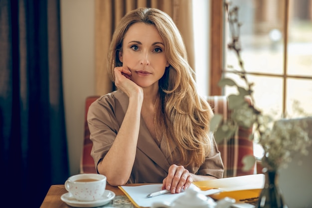 In Gedanken. Blonde langhaarige Frau, die am Tisch sitzt und nachdenklich aussieht