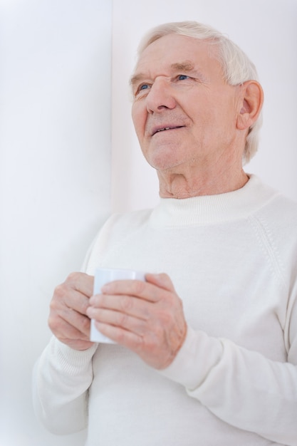 In Gedanken verloren. Nachdenklicher älterer Mann, der eine Tasse Kaffee hält und wegschaut, während er sich an die Wand lehnt