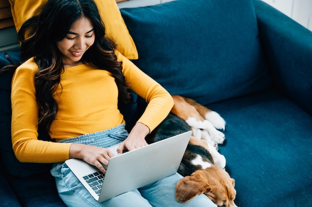 In ihrem Heimbüro auf dem Sofa arbeitet eine Frau fleißig an ihrem Laptop, während ihr Beagle-Hund ein Nickerchen genießt. Ihre Freundschaft und ihr Lächeln definieren diesen gemütlichen Arbeitsplatz.