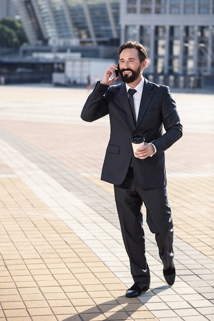 In Kontakt kommen. Fröhlicher Handsoem-Geschäftsmann, der zur Arbeit geht, während er ein Gespräch am Telefon hat