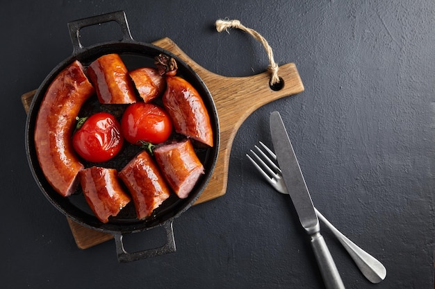 Foto in scheiben geschnittene gebratene schweinswurst in einer gusseisernen pfanne mit tomaten und besteck auf einem holzschneidebrett auf einem dunklen betontisch