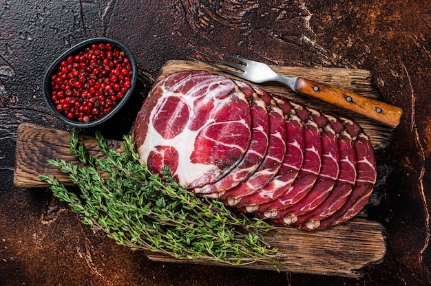 In Scheiben geschnittener Coppa-Schinken auf Holzbrett mit Thymian. Dunkler Hintergrund. Draufsicht.