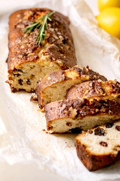 In Scheiben geschnittener hausgemachter Zitronenkuchen mit Rosinen, Nüssen und Rosmarin auf zerknittertem Backpapier, serviert mit frischen Zitronen auf weißem Marmortisch. Nahansicht