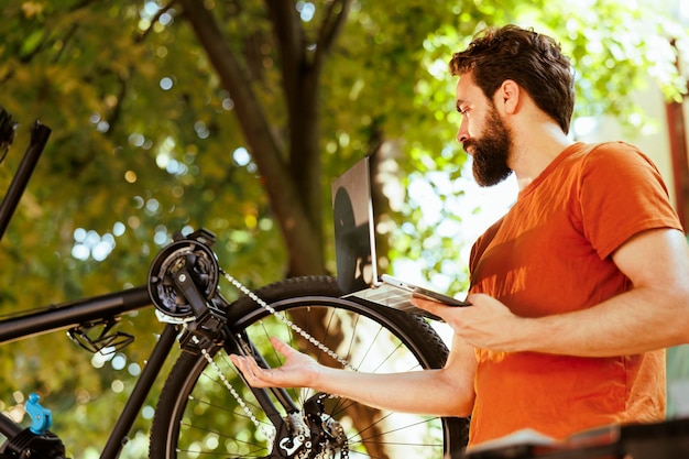 In seinem Garten untersucht ein aktiver, sportbegeisterter Mann mithilfe eines intelligenten digitalen Geräts Möglichkeiten zur Fahrradreparatur. Kaukasischer männlicher Radfahrer inspiziert beschädigtes Fahrrad mit Laptop im Freien.