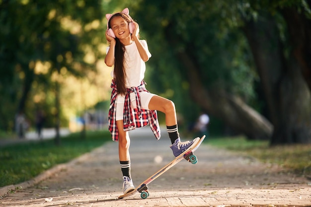 Foto in süßen rosa kopfhörern mit ohren ein glückliches kleines mädchen mit skateboard im freien