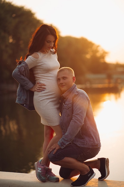 In voller Länge begeisterter Mann in Jeansjacke, der sein zukünftiges Baby im Bauch seiner Frau zuhört und umarmt Sie sind in einem wunderschönen Park am Fluss oder See bei Sonnenuntergang