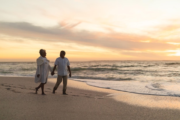 In voller Länge ein älteres, gemischtrassiges Paar, das Händchen haltend am Ufer am Strand spaziert und den Sonnenuntergang genießt