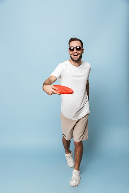 In voller Länge eines fröhlichen aufgeregten Mannes, der ein leeres T-Shirt trägt, das isoliert über der blauen Wand steht und Frisbee spielt