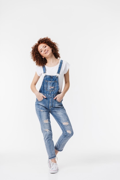 In voller Länge eines fröhlichen jungen rothaarigen, lockigen Mädchens, das Denim-Overalls trägt, die isoliert über weißer Wand posiert?