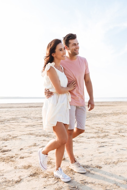 In voller Länge eines schönen lächelnden jungen Paares, das Sommerkleidung trägt, die am Strand spazieren geht und sich umarmt