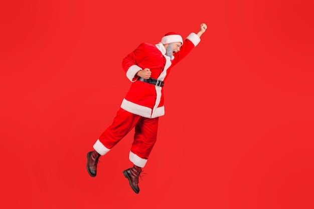 In voller Länge fröhlicher positiver Weihnachtsmann, der hoch springt und wie ein Superheld in der Luft fliegt, in Eile, Winterferien zu feiern. Studioaufnahme im Innenbereich isoliert auf rotem Hintergrund