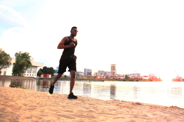 In voller Länge junger afrikanischer Mann in Sportkleidung beim Joggen im Freien, am Strand bei Sonnenuntergang oder Sonnenaufgang.