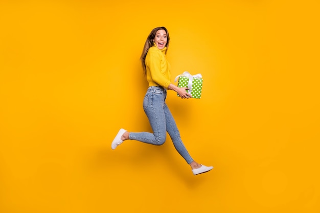 In voller Länge Profil Seite Foto von erstaunt verrückten funky Mädchen wie Urlaub Verkäufe halten grün gepunktete Geschenkbox bringen Freund Sprunglauf tragen Freizeitstil Kleidung isoliert gelbe Farbe Wand