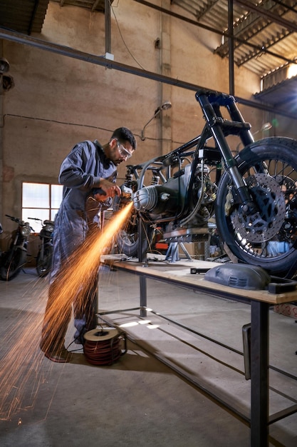 In voller Länge steht ein bärtiger Mann in Uniform mit Schutzbrille an der Werkbank und schneidet mit einer Schleifmaschine in der Garage Metallteile eines Motorrads mit Funkenflug